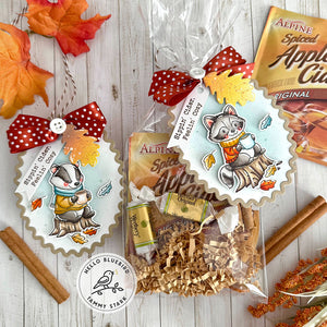 Gift bag with autumn-themed design featuring a racoon and badger, surrounded by cinnamon sticks and leaves on a wooden surface.