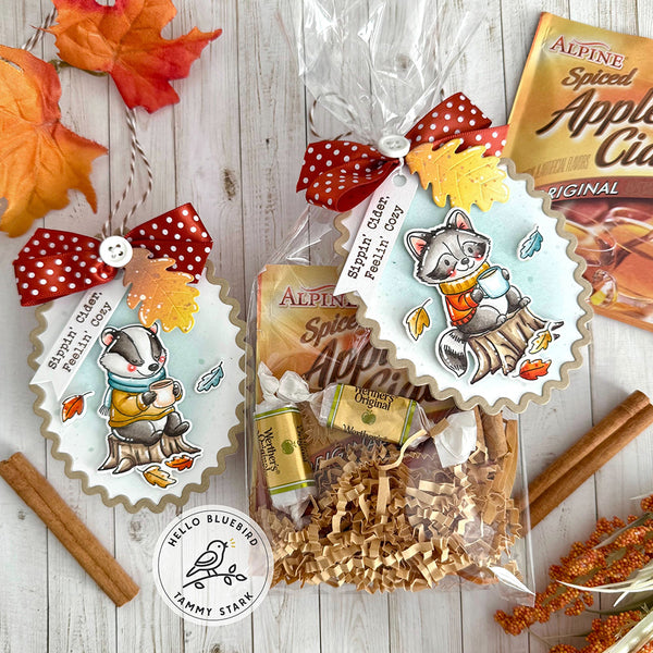 Gift bag with autumn-themed design featuring a cat and dog, surrounded by cinnamon sticks and leaves on a wooden surface.